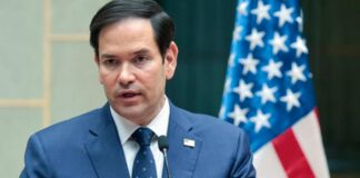 A man in a blue suit speaking into a microphone with an American flag in the background