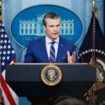 Defense Secretary’s Bold Call: Rules or Camaraderie? Man speaking at podium with flags in background