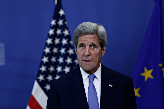 A political figure speaking at a press conference with flags in the background