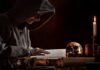 A hooded figure reading an ancient book in a dimly lit room with a skull and candle