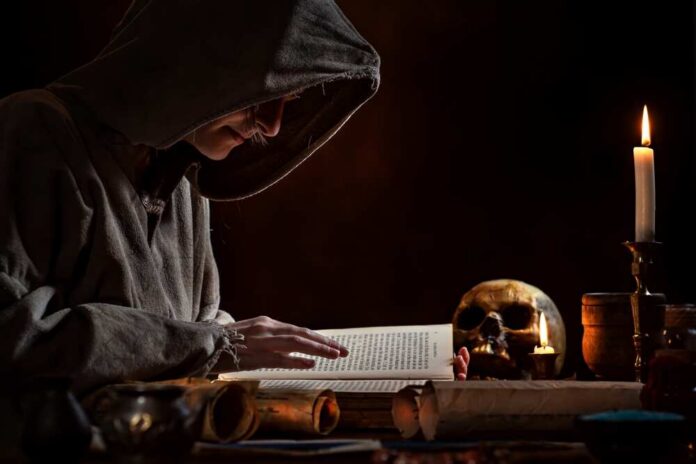 A hooded figure reading an ancient book in a dimly lit room with a skull and candle