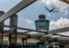 An airplane flying above an airport control tower under a cloudy sky