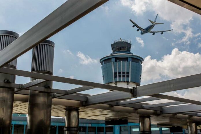 An airplane flying above an airport control tower under a cloudy sky
