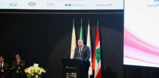 A speaker addressing an audience at a conference with Lebanese flags in the background
