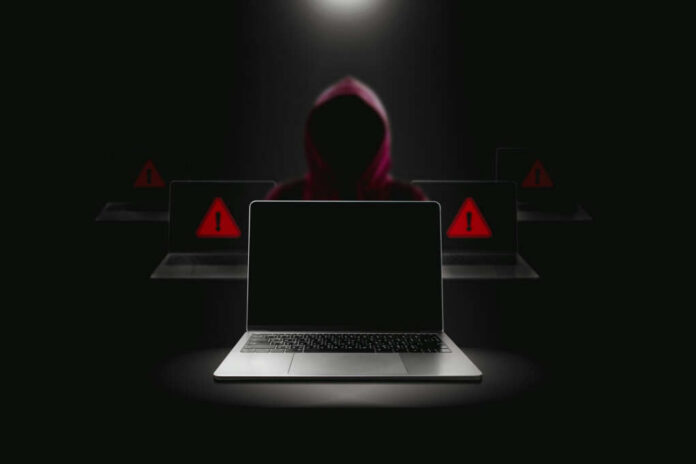 Hacker in hoodie behind laptops with warning signs