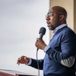 Warnock’s Sermon Sparks Storm Among Evangelicals A man speaking into a microphone at an outdoor event