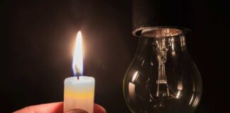 A hand holding a lit candle next to a hanging light bulb