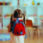A young child with a red backpack standing in a colorful classroom