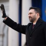 A man in a black coat and gloves waving during a formal event