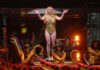A performer in a gold outfit on stage with dancers in the background