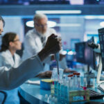 Scientist using pipette at lab bench with test tubes and monitors