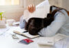 Person with head down on a desk surrounded by paperwork and a calculator