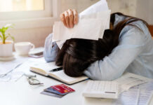 Person with head down on a desk surrounded by paperwork and a calculator