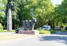 A large 'W' sculpture at a park entrance surrounded by trees