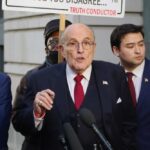 Panic Over Giuliani’s Health—Cause Still Unknown! A man speaking at a press conference with a protest sign in the background