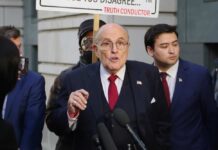 A man speaking at a press conference with a protest sign in the background