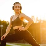 Smiling woman in sportswear wearing headphones, posing outdoors during sunset
