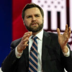 Man in suit speaking on stage with gesturing hands
