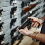 Person holding a handgun in a gun shop with various firearms displayed on the wall