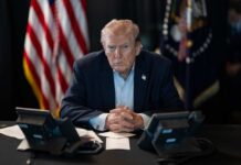 A serious-looking man seated at a table with phones and an American flag in the background