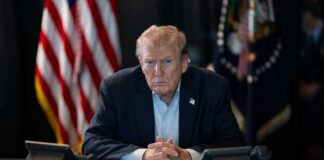 A serious-looking man seated at a table with phones and an American flag in the background
