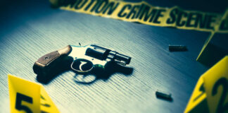 A handgun on a table with evidence markers and crime scene tape in the background
