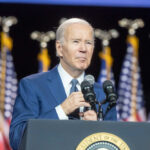 Questions Grew After Biden’s Holiday Message Man speaking at podium with American flags behind.