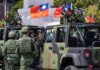 Soldiers beside camouflaged military vehicles with mounted guns and flags