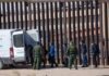 Border agents escort a group near a tall fence and a patrol van