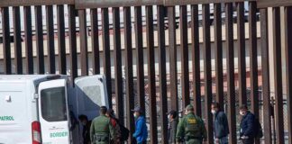 Border agents escort a group near a tall fence and a patrol van