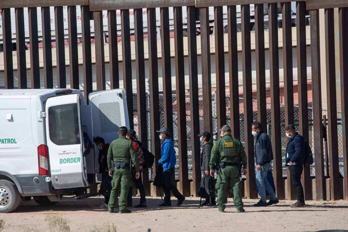 shutterstock_2079408475 Border agents escort a group near a tall fence and a patrol van
