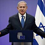 Netanyahu Scraps Tour, Flies In-And-Out man in suit speaking at a podium with Israeli flag
