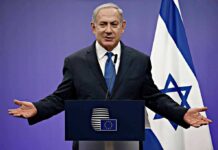 man in suit speaking at a podium with Israeli flag