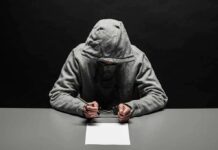Person in gray hoodie writing while handcuffed at a table