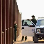 Border patrol agents near a tall fence and a marked pickup truck