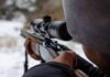person aiming a rifle with a scope outdoors