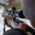 person aiming a rifle with a scope outdoors