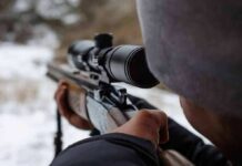person aiming a rifle with a scope outdoors
