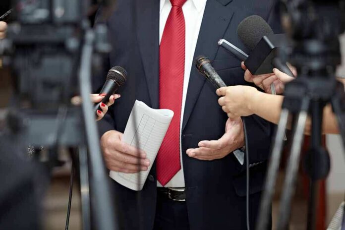 Man in suit with red tie speaking to reporters holding microphones