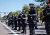 Bear Pascoe, Brian Folkerts, and Eugene Sims among riot police standing in a line on a city street