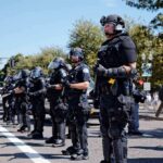 Bear Pascoe, Brian Folkerts, and Eugene Sims among riot police standing in a line on a city street